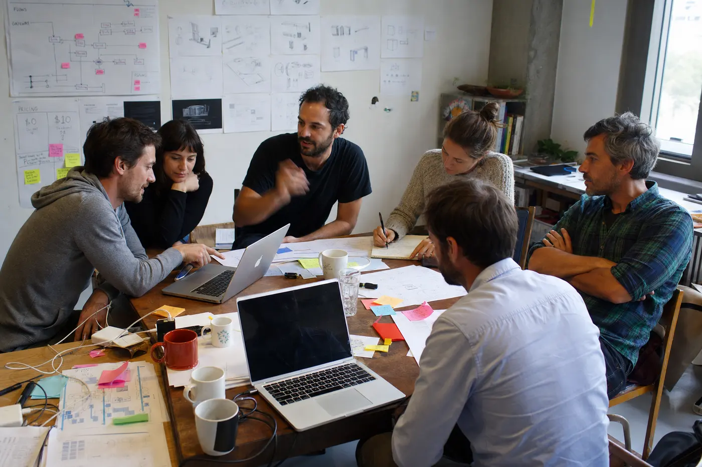 A delivery team in everyday clothes debating a workflow around a table covered in laptops and diagrams